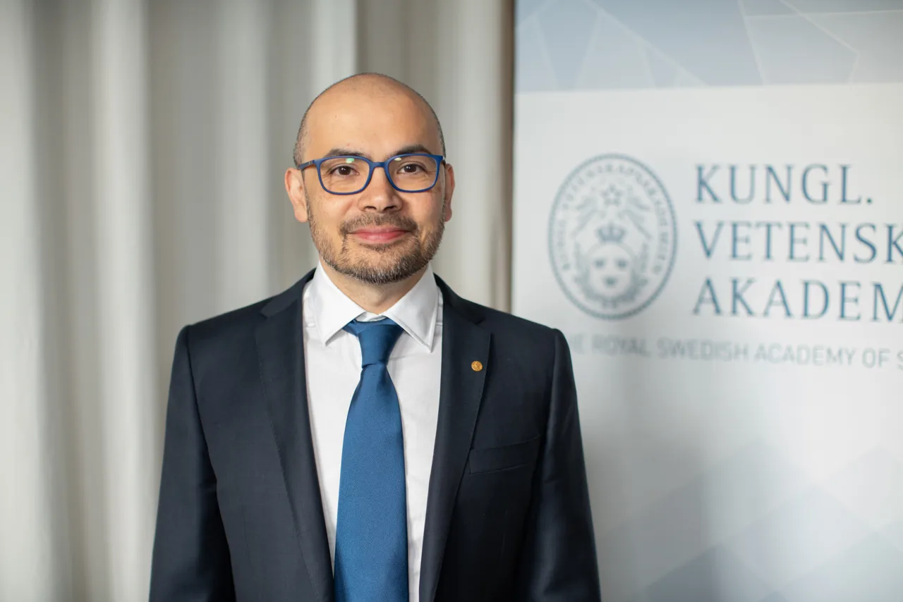 Demis Hassabis con traje y gafas ante el emblema de la Real Academia Sueca de Ciencias.